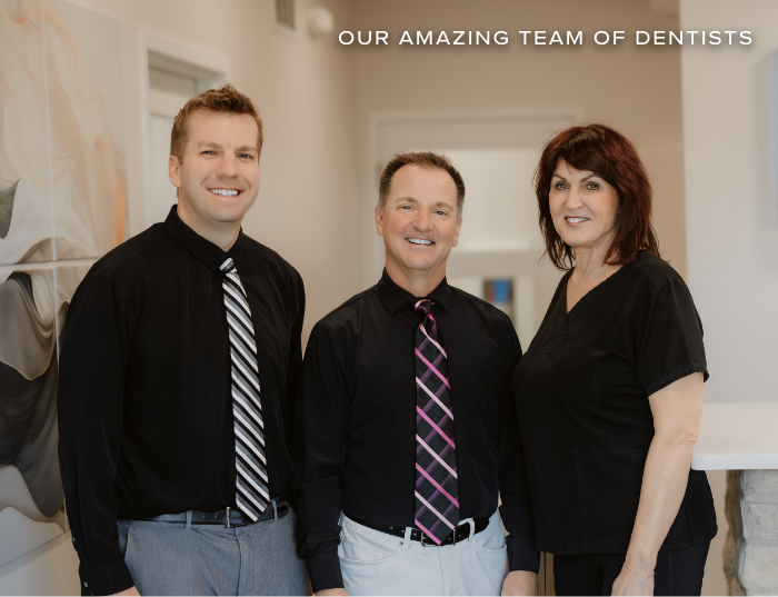 Three smiling dentists in professional attire stand side by side in a modern office, conveying a welcoming and friendly atmosphere. 